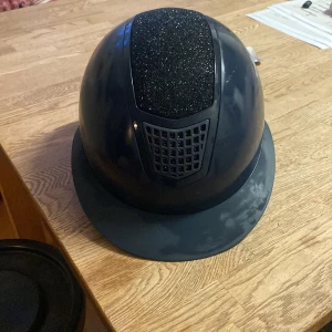 Mörkblå ridhjälm CRW strl S - Snygg  mörkblå ridhjälm från CRW med glittrig panel upptill och ventilerad front. Hjälmen har klassisk form med bred skärm och justerbar passform. Perfekt för dig som vill ha både stil och säkerhet i stallet.