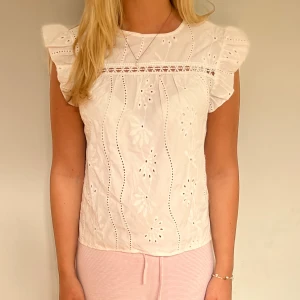 Vit blus med volangärmar och broderi - Superfin vit blus med broderade blommor och hålmönster. Toppen har volangärmar och en söt spetsdetalj över bröstet.