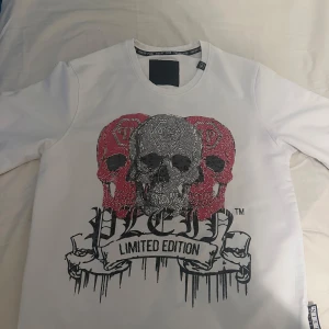 Vit tröja med döskalletryck Philipp Plein - Limited Edition vit tröja från Philipp Plein med stort döskalletryck i svart, silver och rött framtill. Tröjan har rund halsringning, långa ärmar och coola detaljer med glittrande stenar. Perfekt för dig som vill sticka ut med edgy streetstyle.