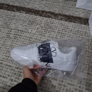 Valentino Garavani vita sneakers - Snygga vita sneakers från Valentino Garavani med bred marinblå rem över vristen och ikoniska nitar på sulan. Skorna har rund tå, platt sula och är tillverkade i skinn. Perfekta för dig som vill ha en clean och lyxig look.