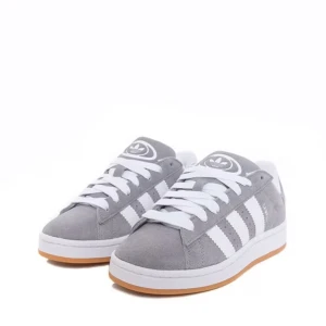 Grå Adidas Campus sneakers mocka - Säljer ett par helt oanvända Snygga grå Adidas Campus sneakers i mocka med klassiska vita ränder och vit sula med gummidetalj. Skorna har rund tå, platt sula och vita skosnören. Perfekta för en clean och sportig stil.