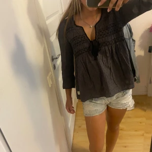 Svart blus med knyt från Noa Noa - Mysig svart blus från Noa Noa med v-ringning och knyt framtill. Blusen har långa ärmar, broderade detaljer och lätt rynkad struktur. Perfekt för dig som gillar boho-vibbar och vill ha något unikt i garderoben.