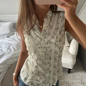 Blommig ärmlös blus med spetsdetaljer - Supersöt ärmlös blus i vit med små blommor i grönt och grått. Blusen har spetsdetaljer längs bröstet och knäppning framtill. Perfekt till jeans för en romantisk och fräsch look. Lätt och luftig, passar perfekt till varmare dagar.