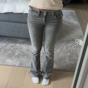 Grå bootcut jeans med fickdetaljer - Säljer ett par grå jeans med bootcut-modell och snygga fickor med knappar bak. Jeansen har låg midja och klassisk femficksdesign. Perfekta för dig som gillar en avslappnad men ändå trendig look.