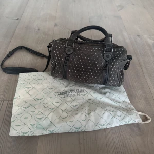 Zadig & Voltaire mocka sunnybag - Cool grå axelväska i mocka från Zadig & Voltaire, täckt med silvriga nitar. Väskan har både handtag och justerbar axelrem samt dragkedja upptill och på sidorna. 
