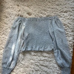 Ljusblå offshoulder blus med volang - Supersöt ljusblå blommig offshoulder blus med smockad kropp och volangkant nertill. Sparsamt använd. Från NLY trend, storlek 36