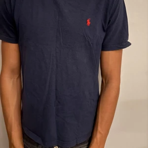 Mörkblå t-shirt från Polo Ralph Lauren - Snygg mörkblå t-shirt från Polo Ralph Lauren med klassisk röd broderad logga på bröstet. Modellen är kortärmad och har en rund hals. Tillverkad i mjuk bomull som känns skön mot huden. Perfekt för dig som gillar stilrena och ikoniska plagg.