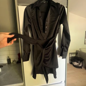 Svart omlottklänning med knytdetalj - Snygg svart kortklänning från Bikbok med omlott-design och medföljande bälte. Klänningen har långärmade ärmar, djup v-ringning med knappar och är gjord i ett glansigt satinliknande material. Perfekt för dig som gillar stilrena och trendiga plagg.