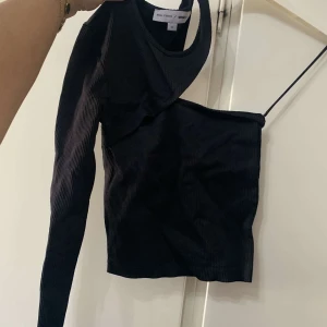 Svart asymmetrisk ribbad topp Gina Tricot - Cool svart ribbad topp från Gina Tricot med unik asymmetrisk design. Ena sidan har lång ärm och andra sidan är ärmlös med smal axelrem. Perfekt för dig som vill sticka ut med en edgy och trendig look.