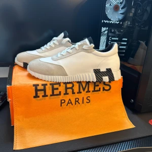 Hermees vita sneakers med mockadetaljer - Exklusiva Hermeea sneakers i vitt skinn med ljusgrå mockadetaljer och svart H-logga på sulan. Snygg ribbad sula och klassisk snörning. Tungan har Hermees Paris-logga och skon har en modern, sportig vibe. Perfekta för dig som gillar lyxiga och stilrena sneakers.