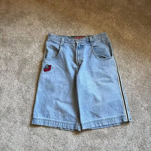 Blå jeansshorts från Skunk 33 - Säljer ett par loose fit blå jeansshorts från Skunk med coolt broderat märke framtill och breda ben. Shortsen har klassiska fickor, dragkedja och bälteshällor. Snygg randig detalj längs sidan och tillverkade i 100% bomull. Perfekt för chill dagar.