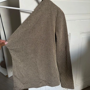Glittrig one shoulder topp från Zara - Säljer en snygg glittrig beige one shoulder topp från Zara. Toppen är ribbad och har lång ärm på ena sidan, vilket ger en cool asymmetrisk look. Perfekt för dig som vill sticka ut lite extra. Materialet är stretchigt och skimrar i ljuset.