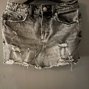 Grå jeanskjol med slitningar från Zara - Snygg grå jeanskjol från Zara med slitna detaljer och rå kant nertill. Kjolen har klassisk femficksdesign och en cool, lite trashad look. Perfekt för dig som gillar streetstyle och vill ha en edgy vibe i garderoben.