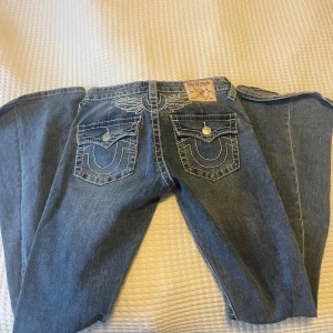 True Religion jeans med broderi, stl 26 - Snygga blå jeans från True Religion med ikoniska vita broderier på bakfickorna och kontrastsömmar. Klassisk femficksmodell bootcut och låg midja. Jeansen har silverfärgade knappar och detaljer som sticker ut.