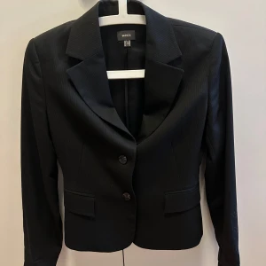 Svart randig kavaj från Mexx - Snygg svart kavaj från Mexx med diskreta tunna ränder. Klassisk modell med två knappar framtill, spetsiga slag och två fickor med lock. Perfekt för dig som gillar stilrena och tidlösa plagg