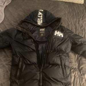 Mörkblå pufferjacka från Helly Hansen - Säljer en svart pufferjacka från Helly Hansen med stor logga på bröstet och huvan. Jackan har dragkedja framtill, två fickor och en rejäl huva. Perfekt för kalla dagar och riktigt snygg streetstil.