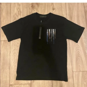 Svart AMIRI t-shirt med färgstänk - Svart t-shirt från AMIRI med stort tryck på både bröstet och ryggen. Trycket består av AMIRI-loggan med coola färgstänk i blått, orange och vitt som rinner ner. Klassisk passform och rund hals. Perfekt för dig som gillar streetwear med edge.