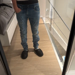 Replay ljusblå slitna jeans herr - Säljer ett par ljusblå jeans från Replay med snygga slitningar och trasiga detaljer framtill. Klassisk femficksmodell med normal passform och raka ben. Jeansen har läderpatch med Replay-logga bak i midjan och är tillverkade i bomull.