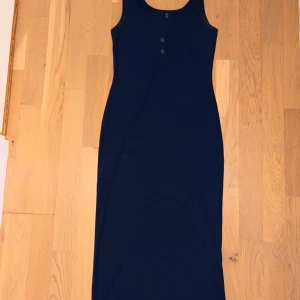 Marinblå ribbad långklänning - Snygg marinblå långklänning med ribbad struktur och rundad hals. Klänningen är ärmlös och har tre knappar framtill för en cool detalj. Perfekt för dig som gillar enkel och stilren design med en touch av sportig vibe.