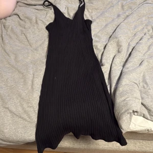 Svart ribbad klänning XS - Svart ribbad klänning med smala axelband och v-ringad hals. Klänningen är figurnära och har en snygg, vågig nederkant. Perfekt för dig som gillar en enkel men stilren look. Materialet är mjukt och stretchigt för en bekväm passform.