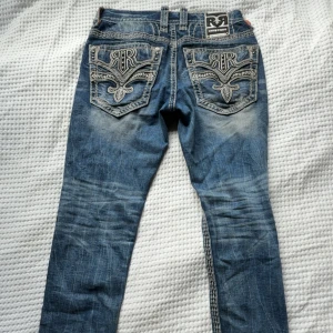 Rock Revival jeans med broderade fickor - Snygga blå Rock Revival jeans med grymma broderade bakfickor och kontrastsömmar. Jeansen har en klassisk straight fit och cool slitning framtill och baktill. Perfekta för dig som vill sticka ut med detaljer och unik stil.