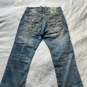 Rock Revival ljusblå jeans med broderi - Säljer ett par ljusblå jeans från Rock Revival med snygga broderade detaljer och silverknappar på bakfickorna. Jeansen har en klassisk femficksdesign, kontrastsömmar och slitna partier för en cool look. Passar dig som gillar y2k och snygga jeans