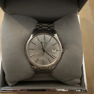 Silverfärgad Calvin Klein herrklocka - Stilren unisexklocka från Calvin Kleini silver  med rund urtavla och datumvisning. Klockan har en klassisk länkarmband i metall med Calvin Klein-logga på spännet. Urtavlan är minimalistisk med tunna svarta markeringar och boetten är också i silverfärgad metall. Fungerar som den ska, batteriet behövs bytas. 
