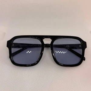 Solglasögon med blå ton - Snygga svarta solglasögon med rektangulär form och blåtonade linser. Bågarna är i blank plast och har en cool retrostil med markerad brygga. Perfekt för dig som vill ha en stilren och klassisk look med en modern twist.