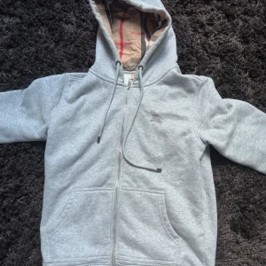 Grå hoodie från Burberry med dragkedja - Säljer en grå hoodie från Burberry med klassiskt rutigt foder i huvan. Tröjan har dragkedja framtill, snörning i huvan och två stora fickor. Tillverkad i mjuk bomull med en broderad logga på bröstet. Perfekt för en chill och stilren look.