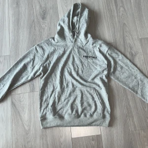 Grå Essentials Fear of God hoodie - Säljer en grå hoodie från Essentials Fear of God med svart logga på bröstet och ryggen. Tröjan har huva, känguruficka och är tillverkad i mjuk bomullsmix. Perfekt för en chill och stilren look.