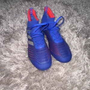 Blåa Adidas Predator fotbollsskor - Säljer ett par blåa Adidas Predator fotbollsskor med röda detaljer. Skorna har snörning, strumpliknande skaft och är tillverkade i syntetmaterial för bästa passform och grepp på planen. Perfekta för dig som vill sticka ut på matchen! Kom privat för mer info!