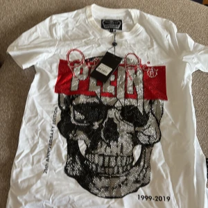 Vit t-shirt med dödskalle från Philipp Plein - Snygg vit t-shirt från Philipp Plein med stor dödskalle framtill, glittriga detaljer och röd text. Märkets logga i nacken och exklusiv Platinum Cut-tag. Perfekt för dig som gillar statement-plagg och cool design.