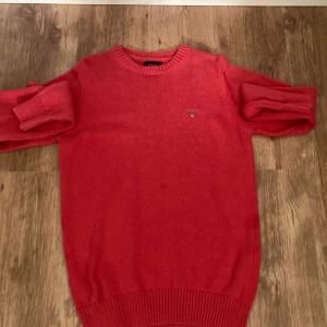 Röd stickad tröja från GANT - Säljer en snygg röd stickad tröja från GANT i storlek S. Tröjan har rund hals, ribbade muddar och ett litet broderat GANT-logo på bröstet. Perfekt för dig som gillar klassisk stil med en färgklick. Materialet är mjukt och skönt stickat.