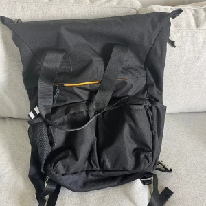 Svart ryggsäck med meshficka - Stor svart ryggsäck med flera fack, dragkedjor och meshficka på sidan. Rymlig design med justerbara axelremmar och extra handtag upptill. Perfekt för skola, träning eller utflykter. Snygg och stilren look med diskreta detaljer och praktisk förvaring.