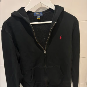 Svart hoodie från Polo Ralph Lauren XL - Svart hoodie från Polo Ralph Lauren i storlek XL. Klassisk design med dragkedja, huva och känguruficka. Liten röd broderad logga på bröstet. Mjuk bomullskänsla, perfekt för chill dagar eller när du vill ha en enkel och stilren look.