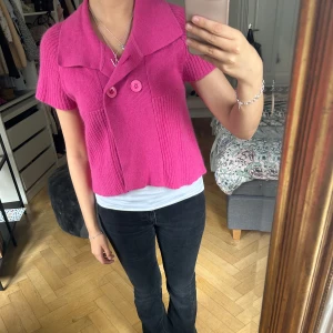 Rosa stickad kofta med stora knappar - Säljer en kort, rosa stickad kofta med stora matchande knappar framtill och bred krage. Koftan har korta ärmar och ribbade detaljer som ger en snygg struktur. Perfekt för dig som vill ha en färgklick i garderoben! 