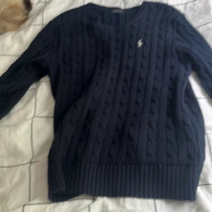Mörkblå stickad tröja Polo Ralph Lauren - Snygg mörkblå stickad tröja från Polo Ralph Lauren med klassisk kabelstickad design och liten broderad logga på bröstet. Tröjan har rund hals och långa ärmar, perfekt för dig som gillar stilrena och tidlösa plagg.