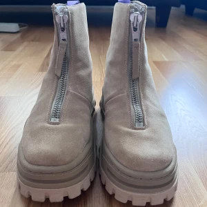 eytys stövlar med dragkedja mocka - Chunky boots från Eytys i beige mocka med grov, mönstrad sula och dragkedja framtill. Skorna har en högre siluett och coola detaljer som ger en edgy vibe. Perfekta för dig som gillar statement-skor och vill sticka ut lite extra. Levereras komplett med två skopåsar i tyg.