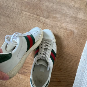 Gucci vita sneakers med röd/grön rand - Säljer ett par äkta Gucci sneakers i vitt skinn med klassiska gröna och röda ränder på sidorna. Skorna har snörning, rund tå och platt sula med Gucci-logga under. Perfekt för dig som vill ha en clean och lyxig look med street-vibe. Va inte rädda för att skriva eller komma med lägre pris