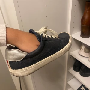 Mörkblå sneakers från Gant i skinn - Snygga mörkblå sneakers från Gant i skinn med vita snören och sula. Silverfärgad detalj på hälen och en randig remsa i rött, vitt och blått baktill. Perfekta för en clean och sportig look.