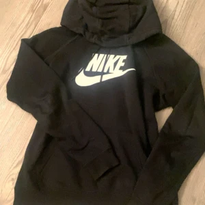 Svart Nike hoodie - Svart hoodie från Nike med stor vit logga framtill. Klassisk modell med huva och känguruficka. Mjuk och skön, passar till det mesta.