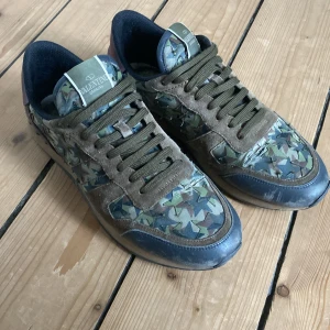 Valentino Rock runners  - Snygga sneakers från Valentino Garavani med camo-mönster i grönt, blått och brunt. Skorna har detaljer i mocka och skinn, samt coola nitar på sulan. Mörkgröna snören och vinröd häl ger extra edge. Perfekta för dig som vill sticka ut med stil. Storlek 43,5. Inga innersulor tyvärr 