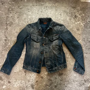 Jeansjacka från Nudie Jeans Co - Säljer en klassisk blå jeansjacka från Nudie Jeans med snygg fade och bronsfärgade knappar. Jackan har två bröstfickor med lock och knappar samt en rak passform. Perfekt för dig som gillar en tidlös och avslappnad stil. Stl s. 