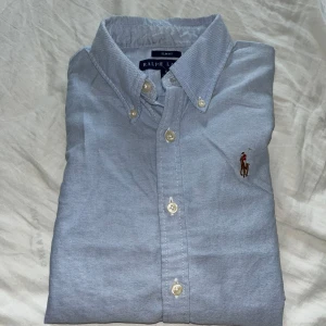 Ljusblå skjorta från Ralph Lauren - Säljer en klassisk ljusblå skjorta från Ralph Lauren i slim fit-modell. Skjortan har långa ärmar, knappar framtill och ikoniska broderade polospelaren på bröstet. Perfekt för dig som gillar stilrena och tidlösa plagg. Skjortan har lite små defekter på vissa ställen så som på sista bilden. Storlek 6/xs-s. Tveka inte att fråga om ni undrar något eller vill ha fler bilder,Mvh Olle 