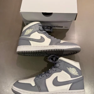 Nike Air Jordan 1 Mid grå/vit 36,5 - Nike Air Jordan 1 Mid i stilren grå och vit färg. Klassisk high-top siluett med snörning och rund tå. Ovandel i skinn med ikoniska Jordan Wings-loggan på sidan. Perfekta sneakers för dig som gillar streetstyle och vill sticka ut med en tidlös modell.