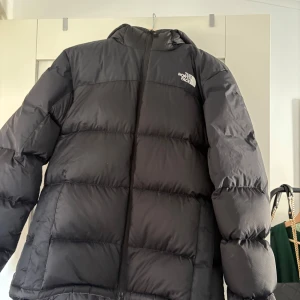 Svart pufferjacka från The North Face - Säljer en svart pufferjacka från The North Face i storlek M. Jackan har en klassisk pufferdesign med huva, dragkedja framtill och logga på bröstet och ryggen. Perfekt för kalla dagar och riktigt skön att bära. Materialet är syntet och dunliknande fyllning (nypris 3500 kr)