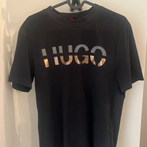 Svart t-shirt från Hugo Boss - Svart t-shirt från Hugo Boss med stor HUGO-logga i silver och guld på bröstet. Klassisk rund hals och korta ärmar. Snygg och stilren modell som passar perfekt till jeans eller shorts.