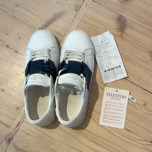 Valentino Garavani vita sneakers - Säljer ett par snygga vita Valentino Garavani sneakers med bred mörkblå rem över vristen och klassisk logga på plösen. Skorna har snörning och är tillverkade i skinn med platt sula. Kommer med originalkartong och tags. Perfekta för dig som gillar exklusiva och stilrena sneakers.ALLT PÅ BILDEN FÖLJS MED. Skriv gärna vid frågor