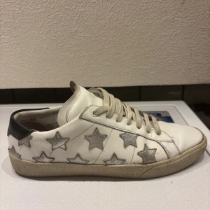 Saint Laurent vita sneakers  - Snygga vita sneakers från Saint Laurent med silverfärgade stjärnor på sidorna och svart detalj vid hälen. Skorna har snörning och är tillverkade i skinn. Perfekta för dig som vill sticka ut med en cool och unik look. Låda och kvitto finns med.