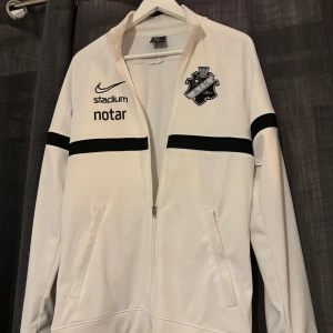 Vit AIK ziptröja från Nike - Säljer en vit ziptröja från Nike med AIK-logga på bröstet och svarta detaljer. Tröjan har tryck från Stadium, notar, Fabege och SVEA, samt svarta ränder över ärmarna. Långärmad med dragkedja och två fickor framtill. Perfekt för dig som älskar fotboll och AIK.